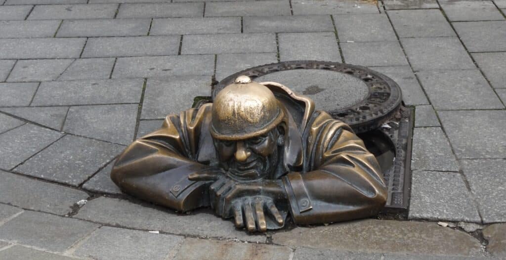 A statue of a man coming out of a vent cover on a cobbled street. He is made of bronze.
