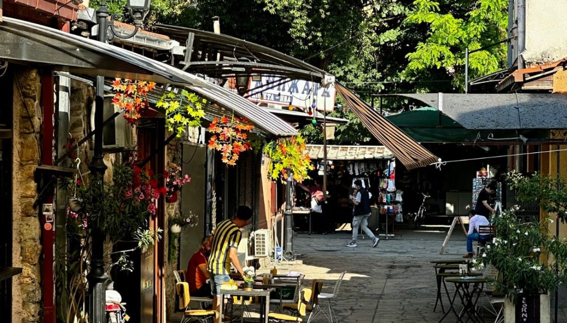 Leafy street with cobbled road. Shops have shaded fronts and it is inviting for people to walk here.