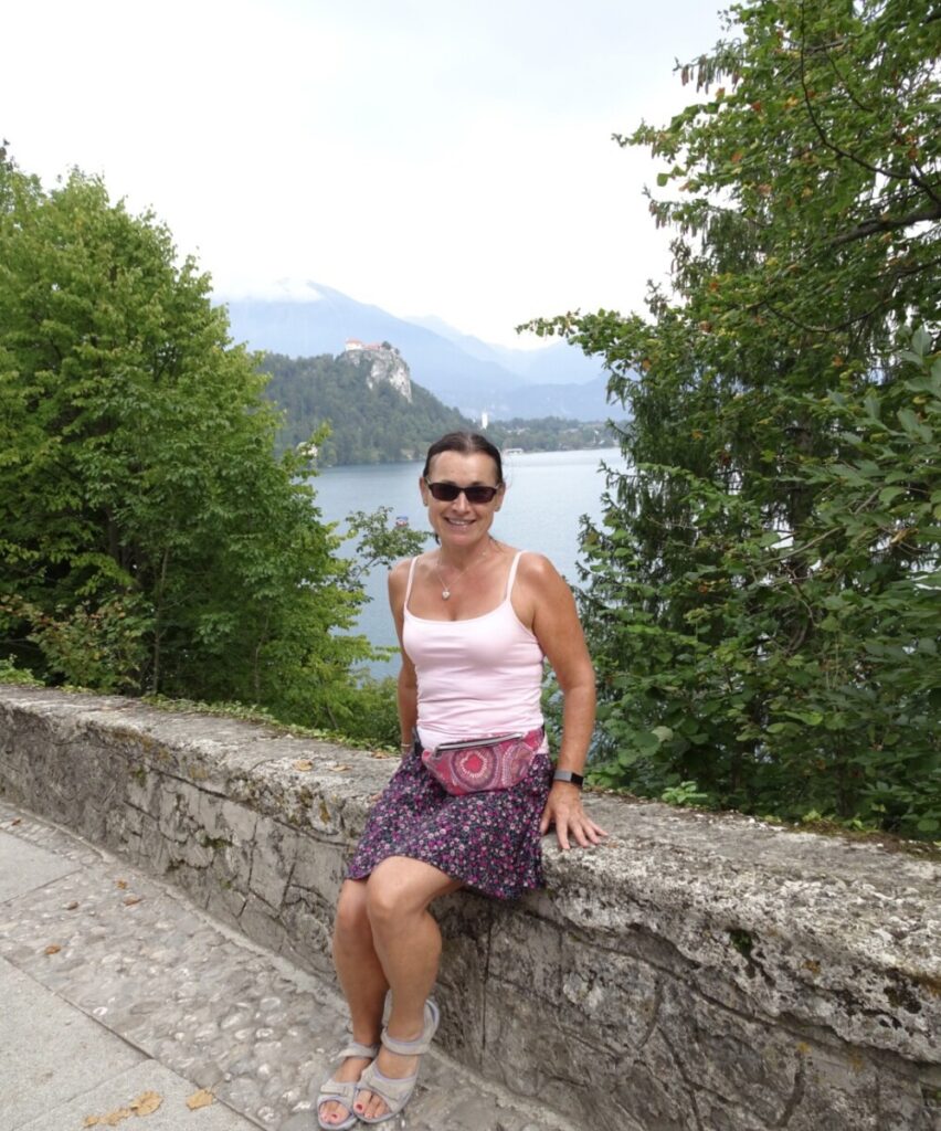 Woman sitting on a stone wall at Lake Bled. Trees surround the lake and mountains are visible in the distance.
