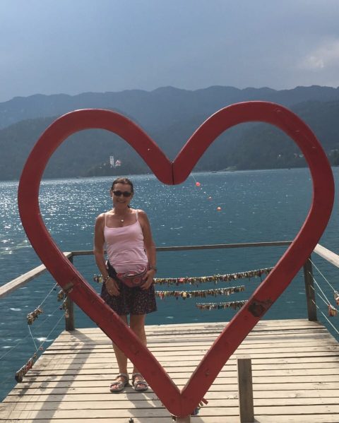 A small pier with wooden boards is secured by a fence. There are many key locks hung onto this fence. A woman stands behind a large red heart, signaling the locks are left in this spot by those in love.