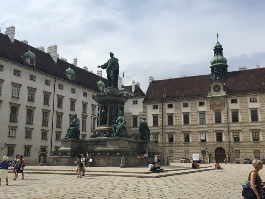 A large square with a statute is framed with ornate multi-story buildings with red tile roofs and towers in Vienna.