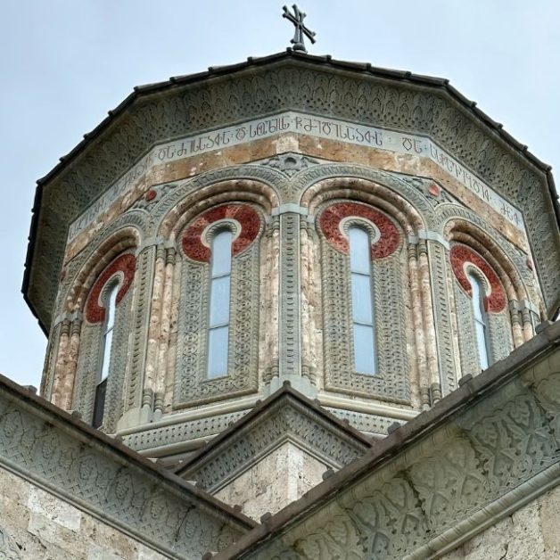 A highly ornate round church steeple with tall rectangular windows rounded at the top with ancient scriptures above and a Georgian cross on top.