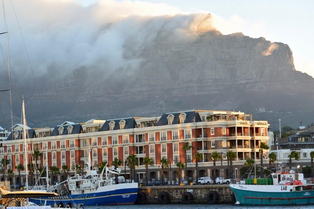 A bay with boats is backed by buildings with brown and yellow coloring. Behind them is a mountain with cloud.