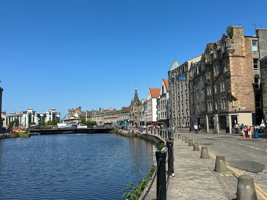 A wide walkway along side waters of Leith near Edinburgh. Old commercial shipping warehouses can be seen along the street, now converted into restaurants and homes.