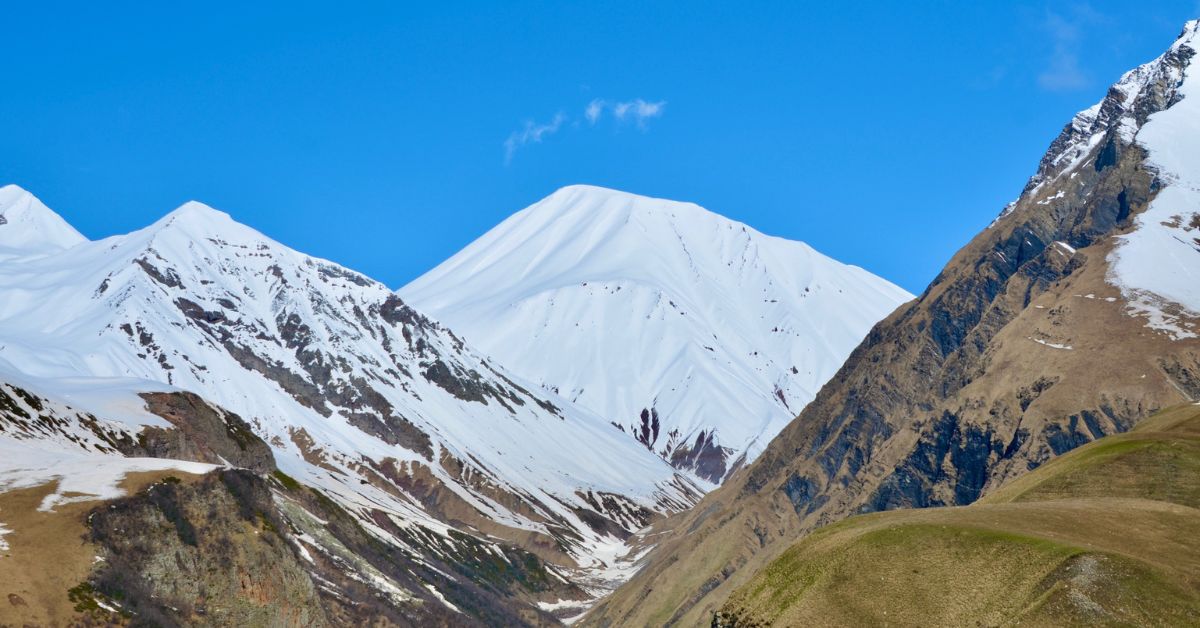View of snow capped peaks and rock faces at lower levels, some with grazing grass on steep slopes.