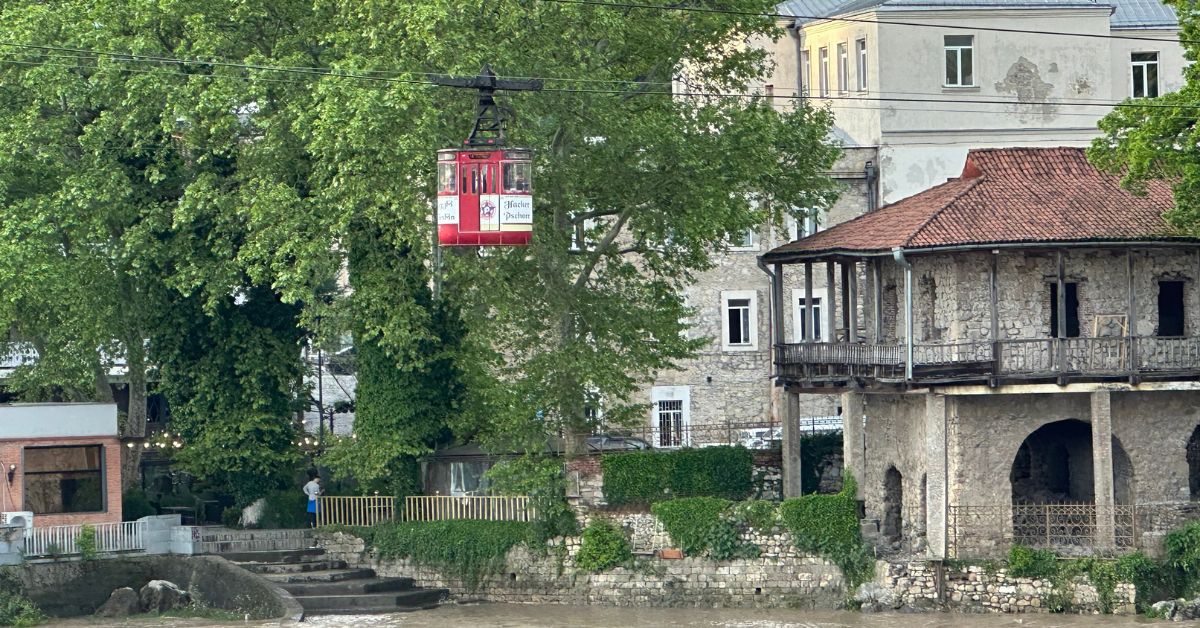 A small cable car in red travels along a track above a river. A stone building is on the right with a red tile roof. Steps lead to the river.