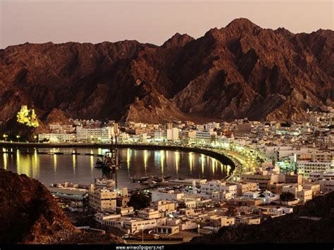 Muscat at night with bay, mountains behind the city, and buildings light up the water.