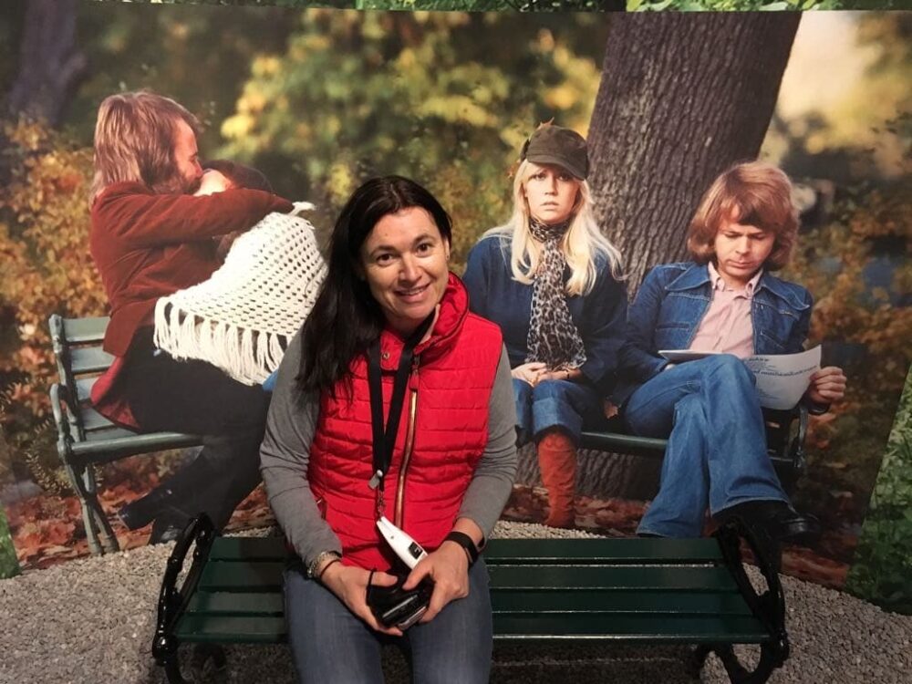 Ruby (co-founder of Two Passports One Planet blog) sitting in Stockholm Sweden on a bench at ABBA museum. Photo of the bands album cover is behind her. Sweden is a great destination to go to July to September.