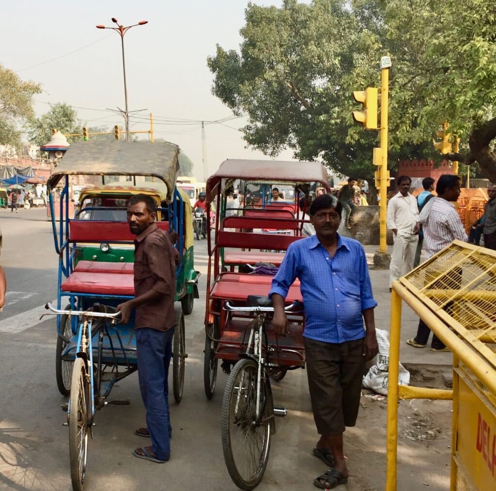 Two cyclists with rickshaws wait for passengers. The small rickshaws have red seats and a sun shade and have space for 2 people.