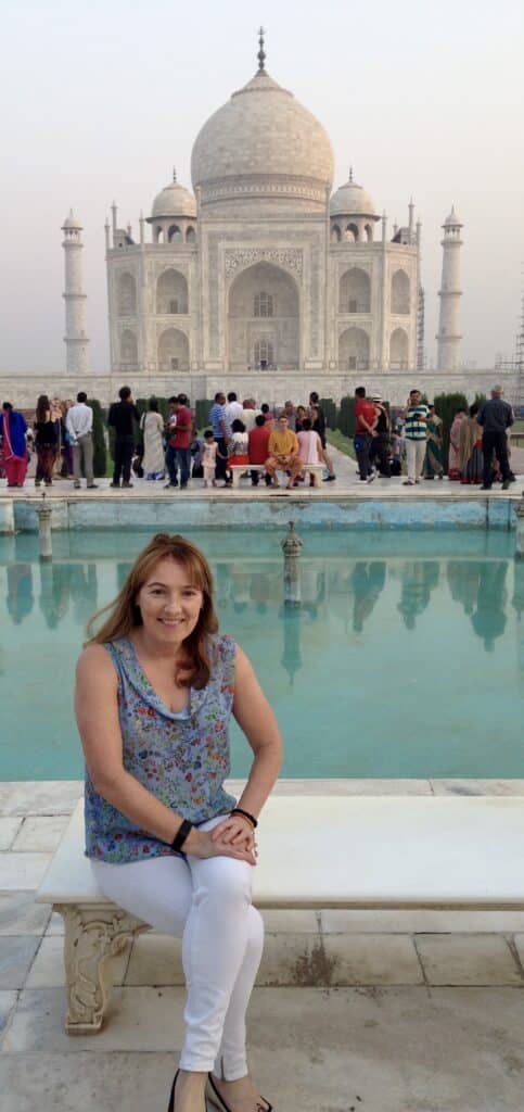 Woman sitting on a bench in front of the Taj Mahal in Agra India. Between her and the building is a pond with reflections of people who are looking at the memorial