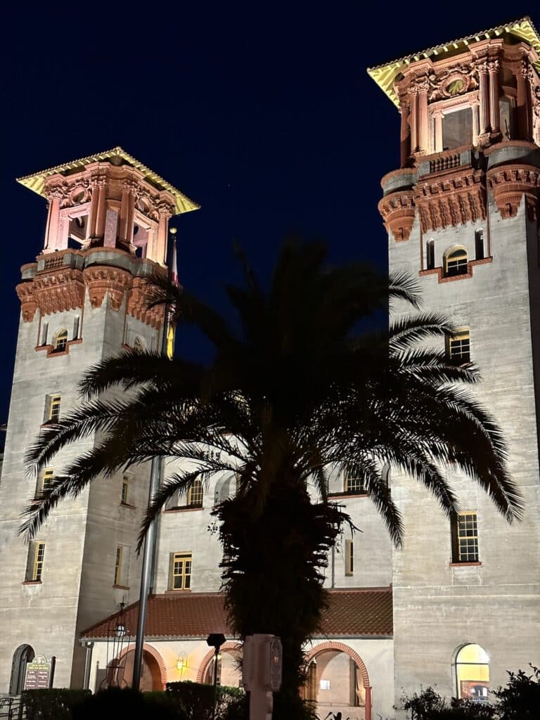 St Augustine - Small Town Destination in Florida. This is a museum lit up at night with a palm tree in front. The building is red and white brick.