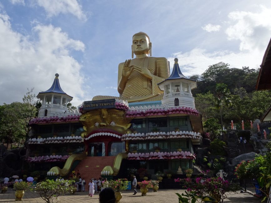 A large golden Buddha stands tall above the ornate Golden temple of Dambulla. It is highly decorated in pink, white and red. The stairs to the temple resemble the feet of a lion.