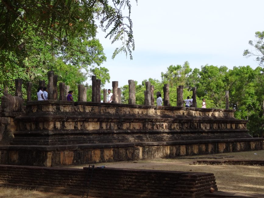 An ancient temple with just the base and pillars now visible. Tourists are exploring the site which is surrounded by trees in full leaf.