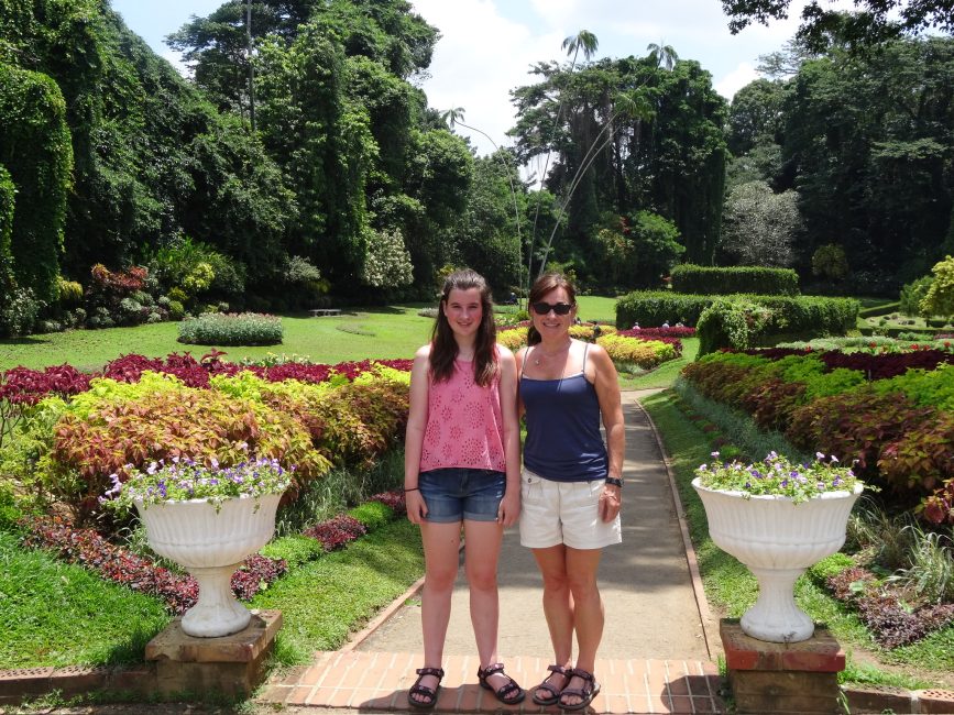 Two women visiting the Kandy Royal Botanic Gardens. They are standing on a pathway surrounded by shrubs, flowers and trees with grass in between.