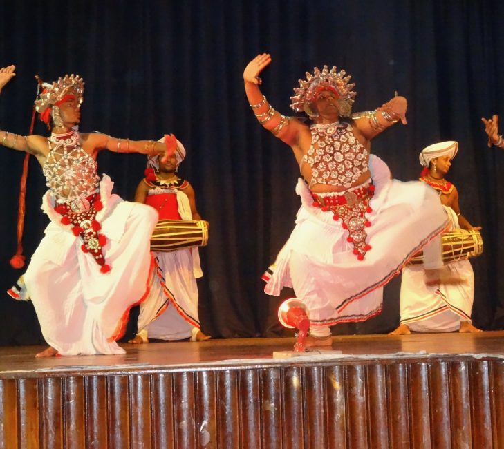 Traditional Sri Lankan dancers on stage. They are wearing white skirt type Tunics and jewel-studded tops and headpieces. Two are playing drums in the background.
