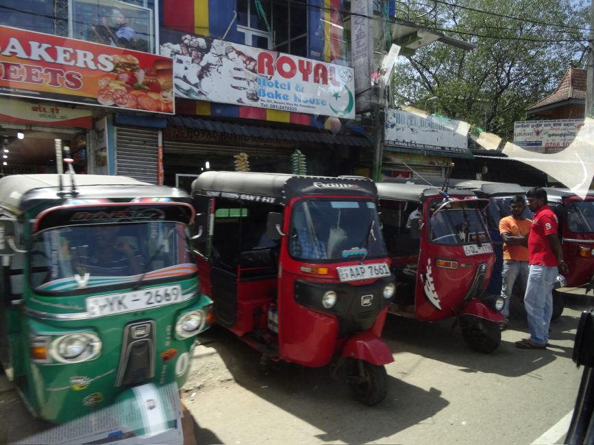 Red and green Tuk Tuk taxis and their drivers are parked waiting for passengers.