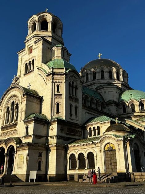 Green and gold domed cathedral in Sofia with people standing at the steps of the church next to a large wooden door.