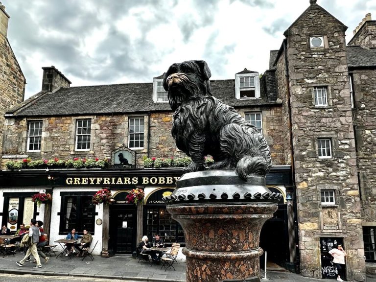 A state of a dog and behind it a stone building and pub called Greyfriars Bobby in Edinburgh.