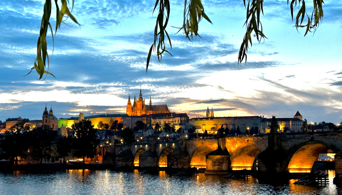 Night time view of Charles Bridge with lights shining on the river below and castle on the hill behind.