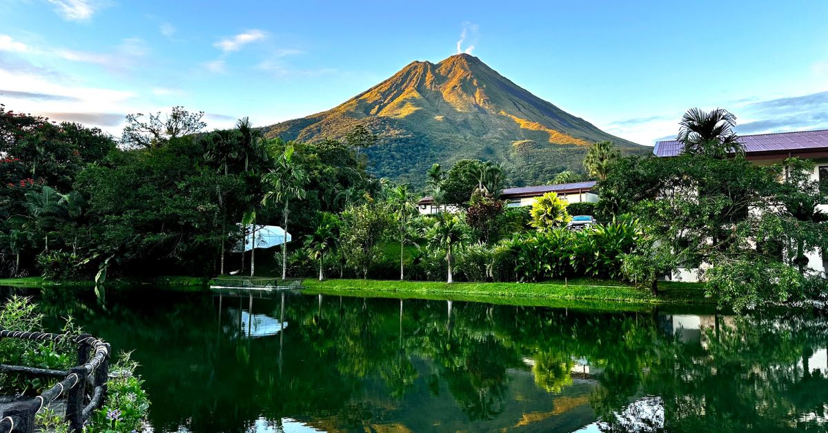 Arenal Volcano with light puffs of smoke coming from the top. In front of volcano is a lake where volcano is reflected in.