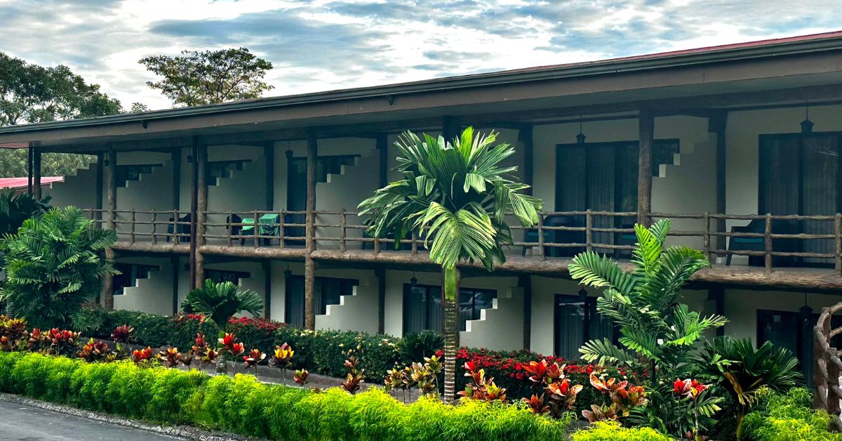 Hotel rooms in a two storey block with tropical plants in front of the rooms