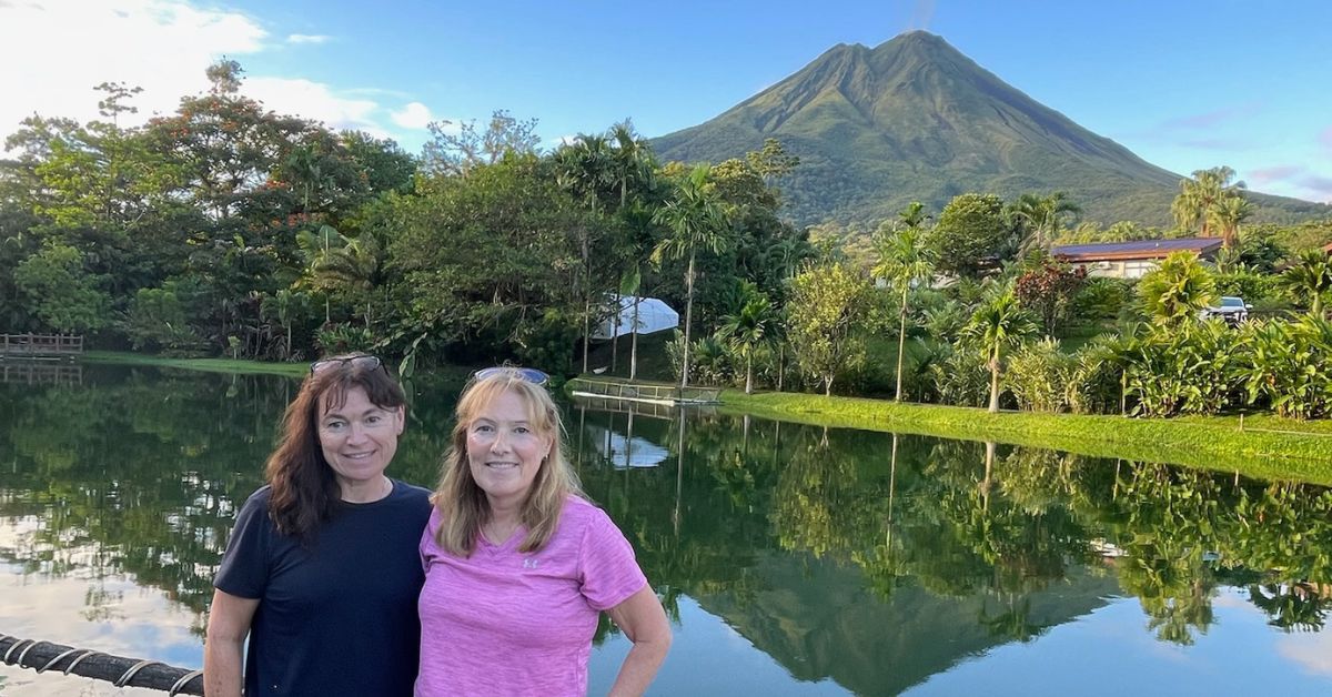 Two women stand infront of a lake. Arenal Volcano is in the background