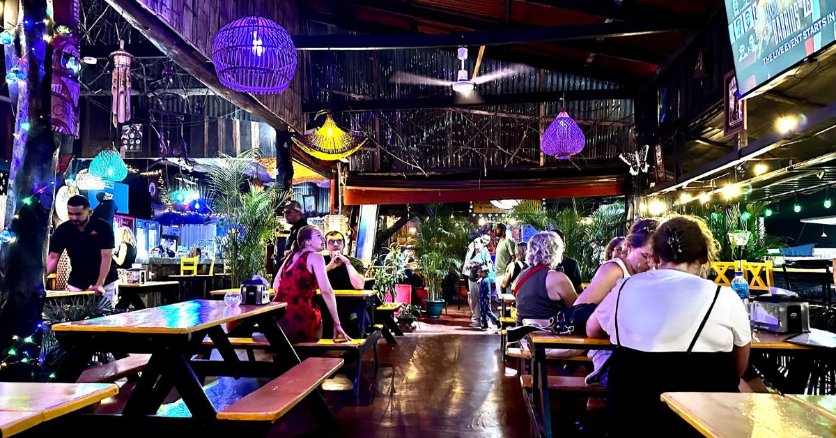 Street food Restaurant with wooden outdoor bench like tables and people sitting down at them.