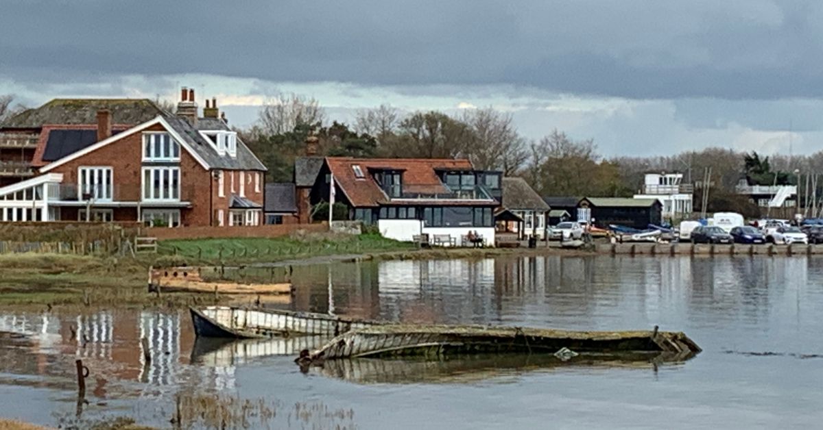 View of Orford Quay with old boats in the foreground and cars parked at the quay in the background. Houses are reflected in the water.