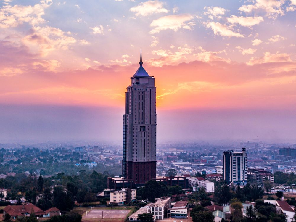 A large tower in central Nairobi surrounded by city buildings. Sunset behind is purpose and pink with clouds.