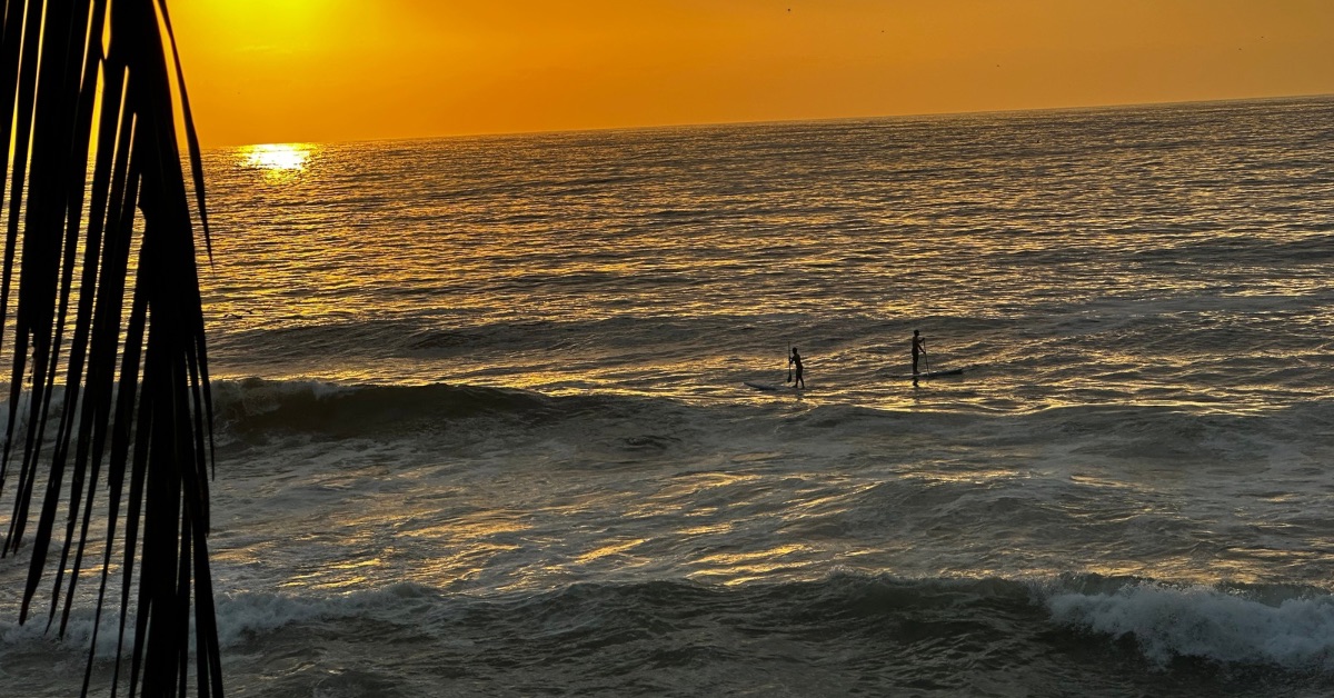 Sun setting on the water while 2 people surfing towards the shore.