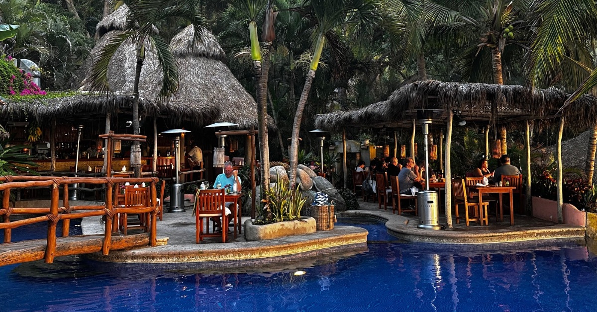 People sitting at tables around a pool area eating under thatch roofed restaurant.