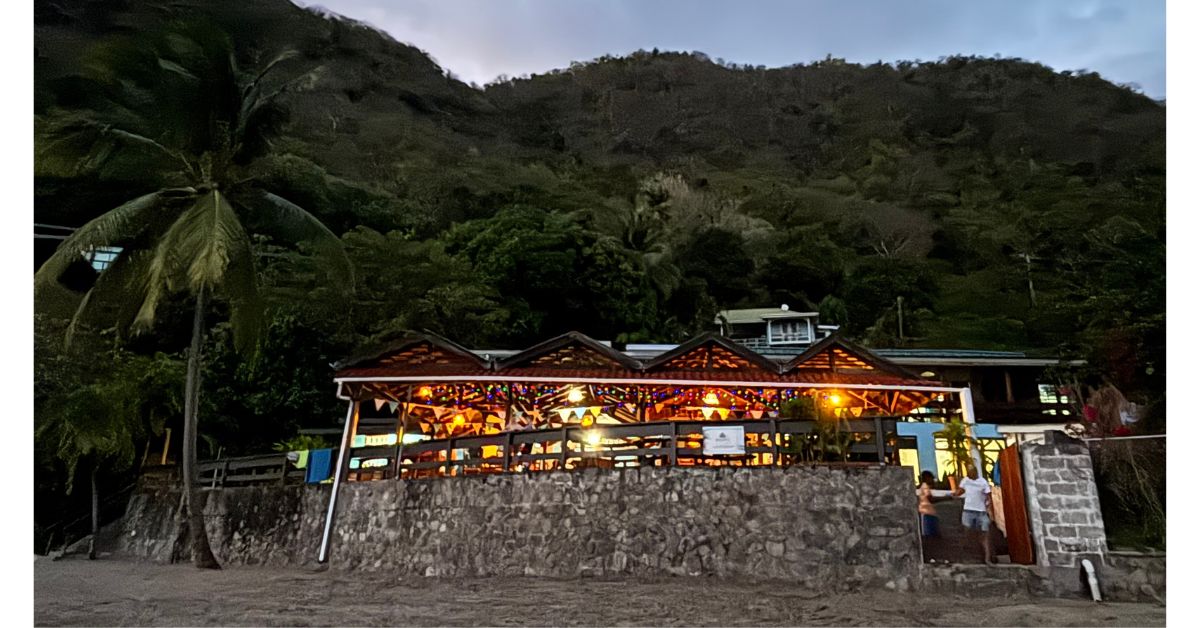 Restaurant at night on the beach lit up with palm trees behind and stone wall in front.