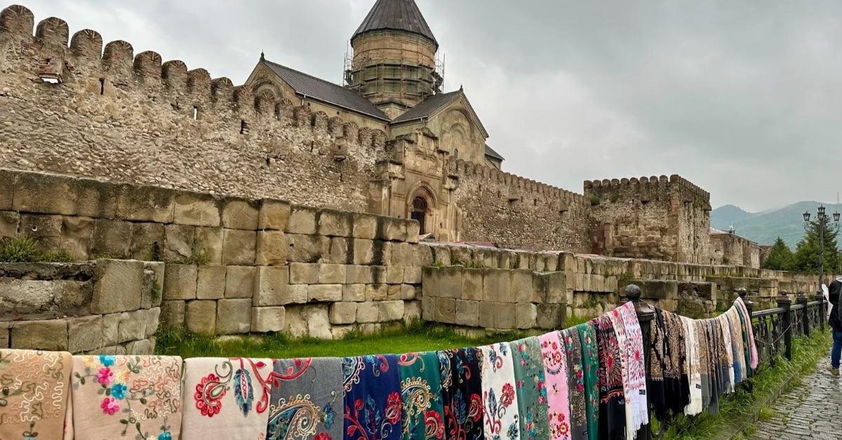 An ancient church in Georgia with an outer wall on which traditional blankets are laid out for sale.