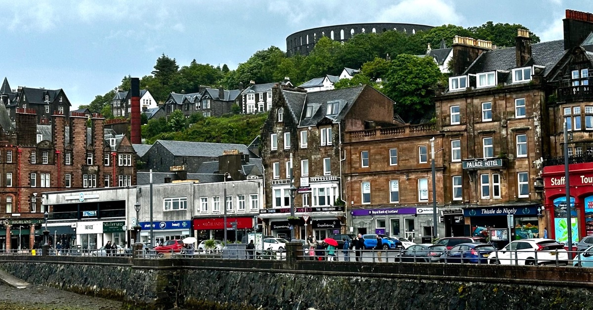 A view of the Main Street of Oban, with red brick multi-story buildings, shops, restaurants and beyond a round stone tower with trees around it.