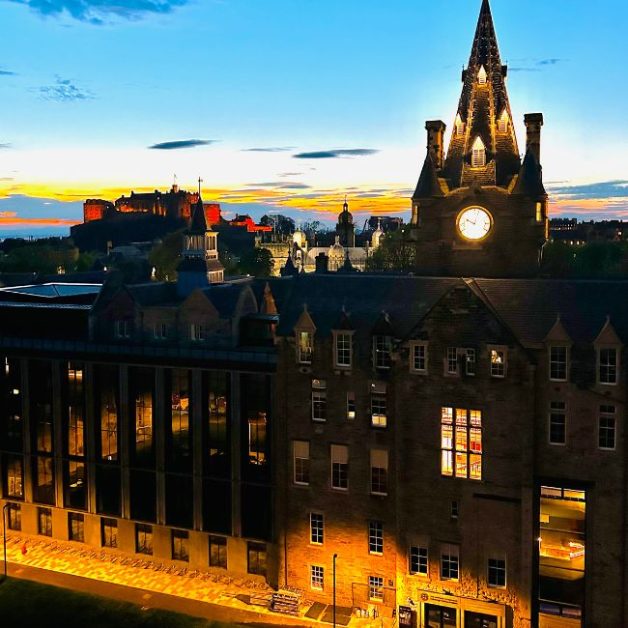 Edinburgh skyline at night over Old Town shows the castle lit up and a gothic steeple clock.